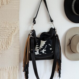 Puede incluir: Un bolso de hombro de cuero negro con un panel central de patr&oacute;n geom&eacute;trico en blanco y negro. El bolso incluye largos flecos en capas que alternan colores negro y beige a ambos lados, sujetos con herrajes de metal en tono plateado.