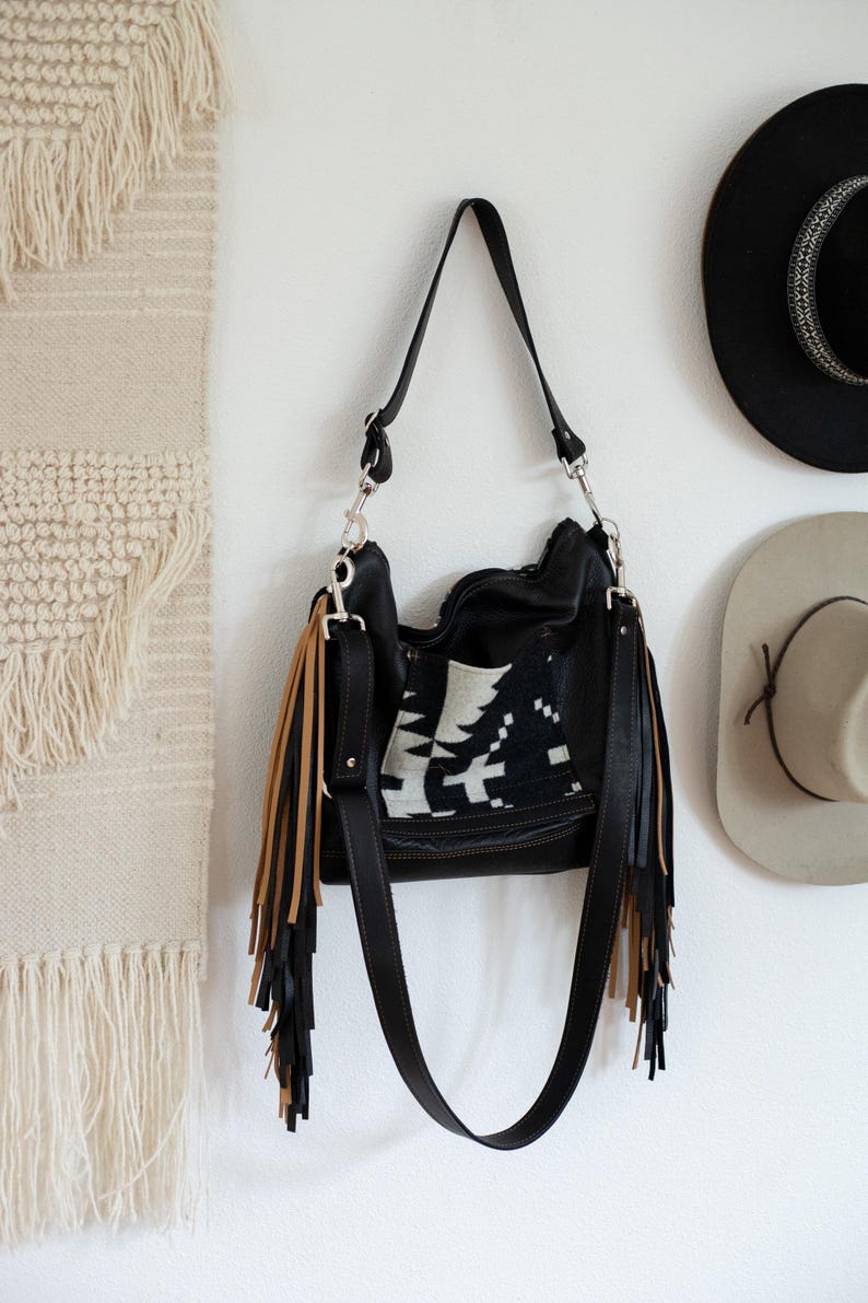 Puede incluir: Un bolso de hombro de cuero negro con un panel central de patr&oacute;n geom&eacute;trico en blanco y negro. El bolso incluye largos flecos en capas que alternan colores negro y beige a ambos lados, sujetos con herrajes de metal en tono plateado.