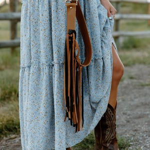May include: A light blue tiered maxi dress with a small yellow floral pattern, paired with brown leather cowboy boots and a brown leather bag strap featuring long, dangling black and brown fringe accents attached with a silver metal clip.