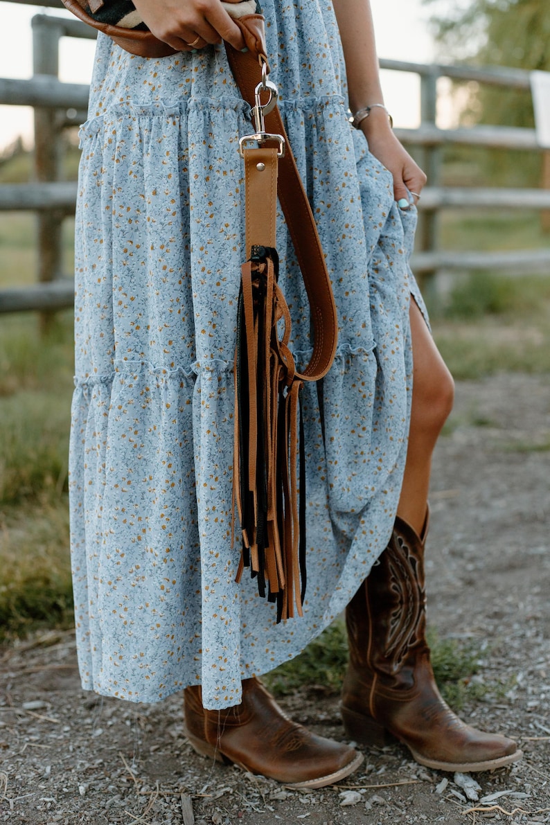 May include: A light blue tiered maxi dress with a small yellow floral pattern, paired with brown leather cowboy boots and a brown leather bag strap featuring long, dangling black and brown fringe accents attached with a silver metal clip.