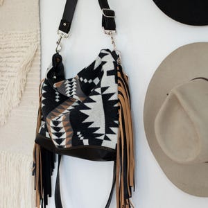 Puede incluir: Un bolso tipo hobo con un patr&oacute;n geom&eacute;trico en negro, blanco y tostado, con largos flecos negros y tostados en los laterales. El bolso incluye una correa de hombro negra ajustable con herrajes de metal plateado y una base de color negro s&oacute;lido.