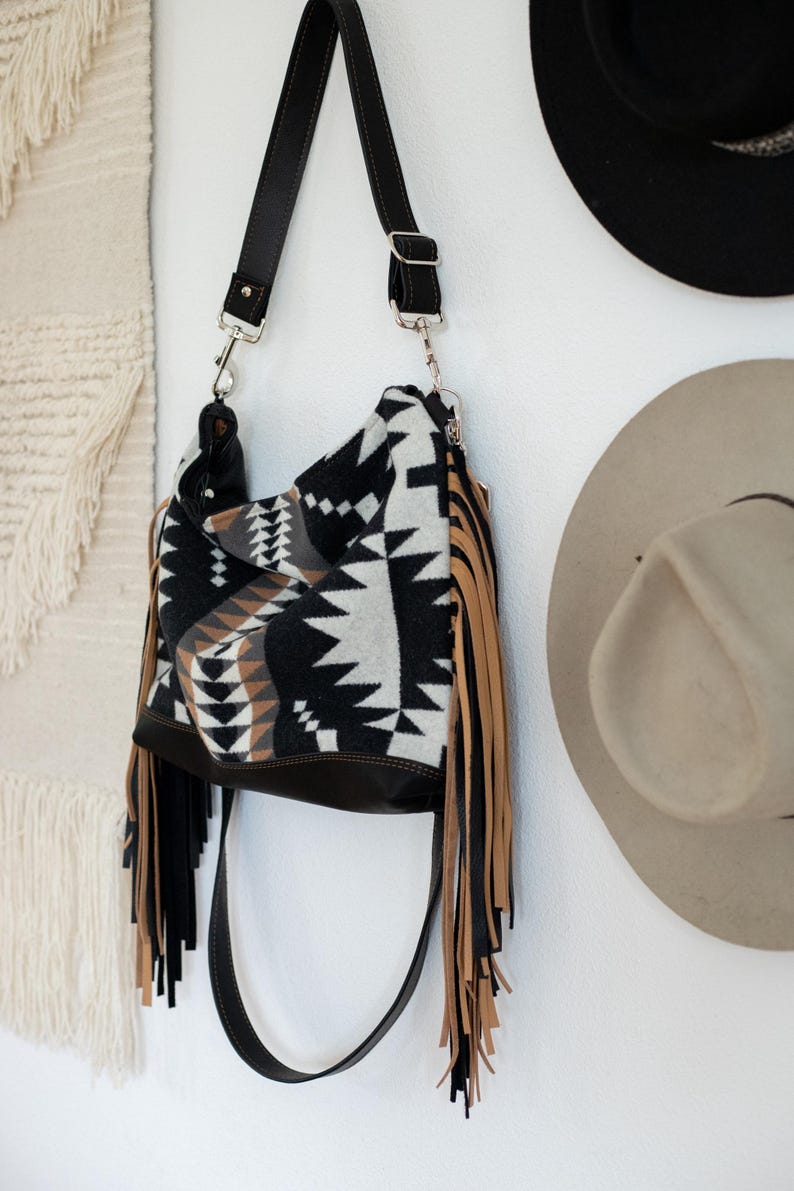 Puede incluir: Un bolso tipo hobo con un patr&oacute;n geom&eacute;trico en negro, blanco y tostado, con largos flecos negros y tostados en los laterales. El bolso incluye una correa de hombro negra ajustable con herrajes de metal plateado y una base de color negro s&oacute;lido.