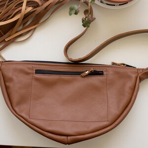 May include: A tan leather fanny pack featuring a crescent shape, a black zippered exterior pocket, and a long adjustable strap. The bag is accented with a large cluster of long, thin tan leather fringe tassels attached to one side with a metal clip.