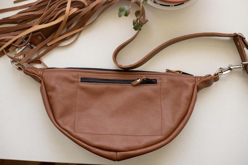 May include: A tan leather fanny pack featuring a crescent shape, a black zippered exterior pocket, and a long adjustable strap. The bag is accented with a large cluster of long, thin tan leather fringe tassels attached to one side with a metal clip.