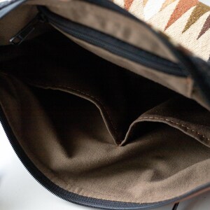 May include: The interior of a shoulder bag featuring a brown fabric lining with two open pockets. The bag has a black zipper closure and an exterior flap with a geometric pattern in shades of cream, black, brown, and olive green, plus a leather strap.
