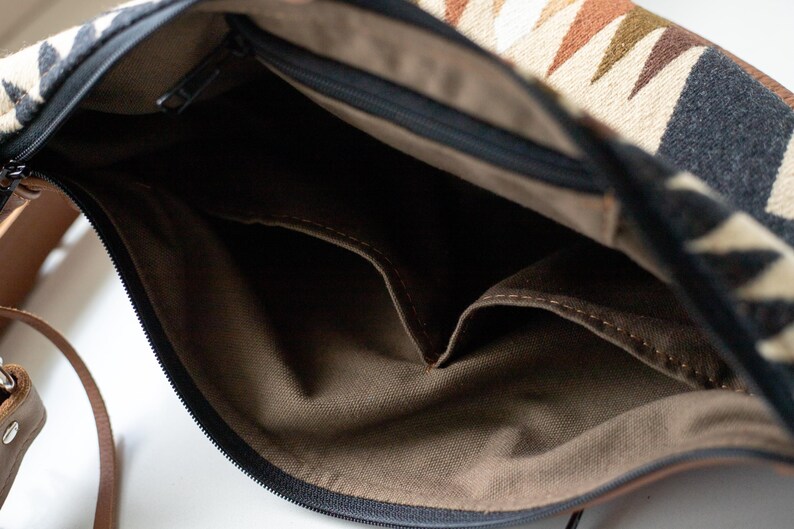 May include: The interior of a shoulder bag featuring a brown fabric lining with two open pockets. The bag has a black zipper closure and an exterior flap with a geometric pattern in shades of cream, black, brown, and olive green, plus a leather strap.