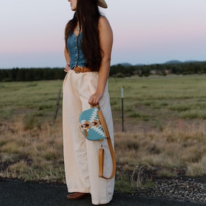May include: A light blue denim vest, cream-colored wide-leg trousers, a tan leather belt, and a cream-colored cowboy hat. A half-moon shaped crossbody bag features a turquoise, white, and brown geometric pattern with a tan leather strap.