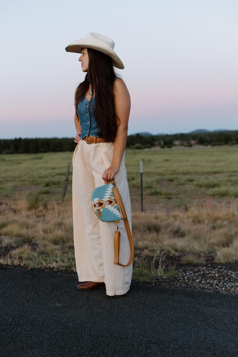 May include: A light blue denim vest, cream-colored wide-leg trousers, a tan leather belt, and a cream-colored cowboy hat. A half-moon shaped crossbody bag features a turquoise, white, and brown geometric pattern with a tan leather strap.