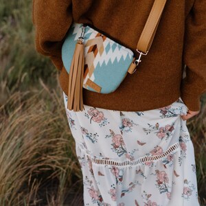 May include: A crossbody bag featuring a light blue, white, and brown geometric pattern, a long tan leather tassel, and an adjustable tan strap. The bag is worn over a brown sweater and a white skirt with a pink and green floral print pattern.