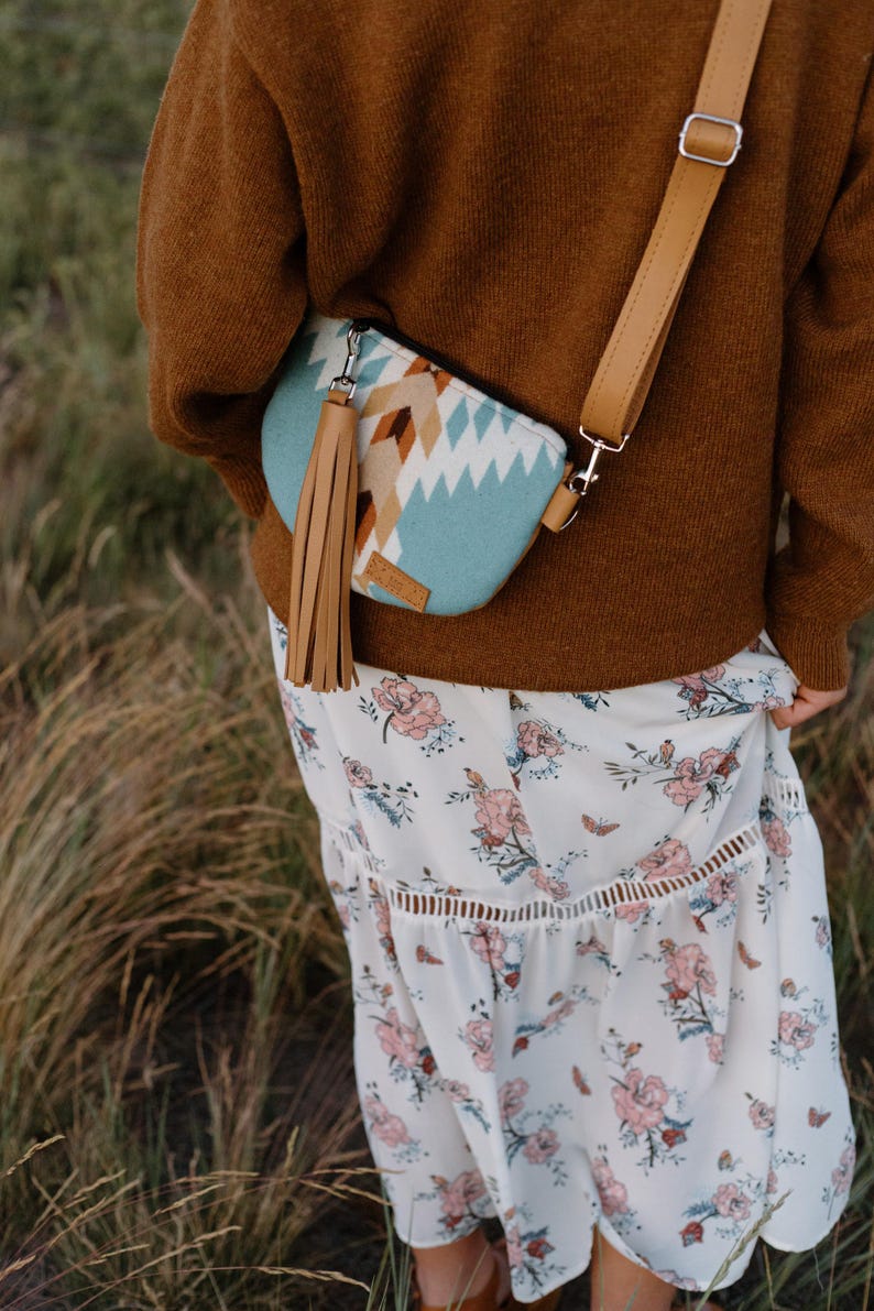 May include: A crossbody bag featuring a light blue, white, and brown geometric pattern, a long tan leather tassel, and an adjustable tan strap. The bag is worn over a brown sweater and a white skirt with a pink and green floral print pattern.