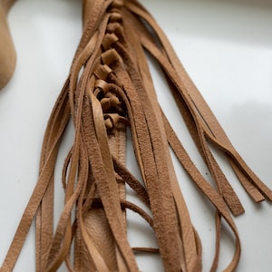 May include: A tan leather strap featuring a braided section that transitions into multiple long, loose fringe strips. The leather has a natural, textured finish and is attached to a silver-toned metal hardware clip at the bottom end of the piece.