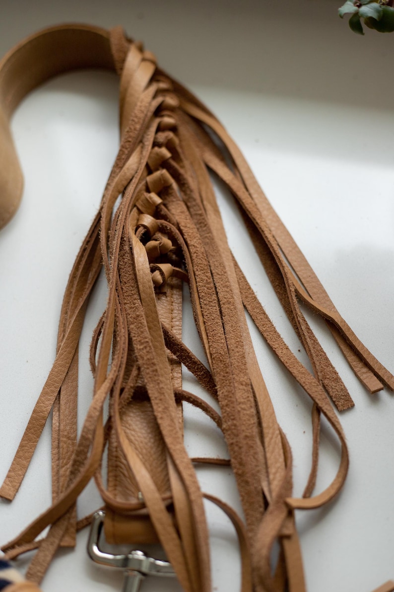 May include: A tan leather strap featuring a braided section that transitions into multiple long, loose fringe strips. The leather has a natural, textured finish and is attached to a silver-toned metal hardware clip at the bottom end of the piece.