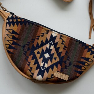 May include: A crescent-shaped crossbody bag featuring a woven fabric panel with a geometric pattern in navy blue, tan, and brown. The bag has tan leather-like trim, a black zipper closure, silver-tone metal hardware, and long tan leather fringe.