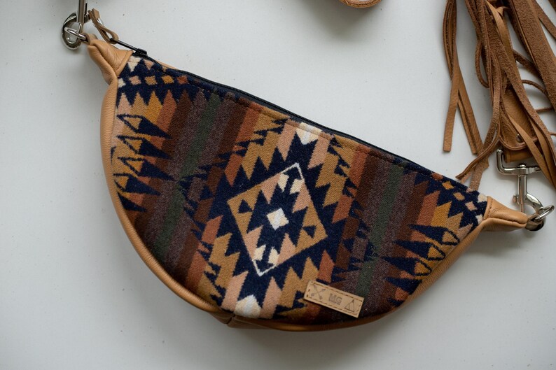 May include: A crescent-shaped crossbody bag featuring a woven fabric panel with a geometric pattern in navy blue, tan, and brown. The bag has tan leather-like trim, a black zipper closure, silver-tone metal hardware, and long tan leather fringe.