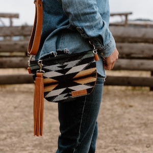 May include: A black, white, and tan patterned crossbody bag with a brown leather strap and tassel. The bag is worn over a denim shirt and jeans. The bag has a small leather tag.