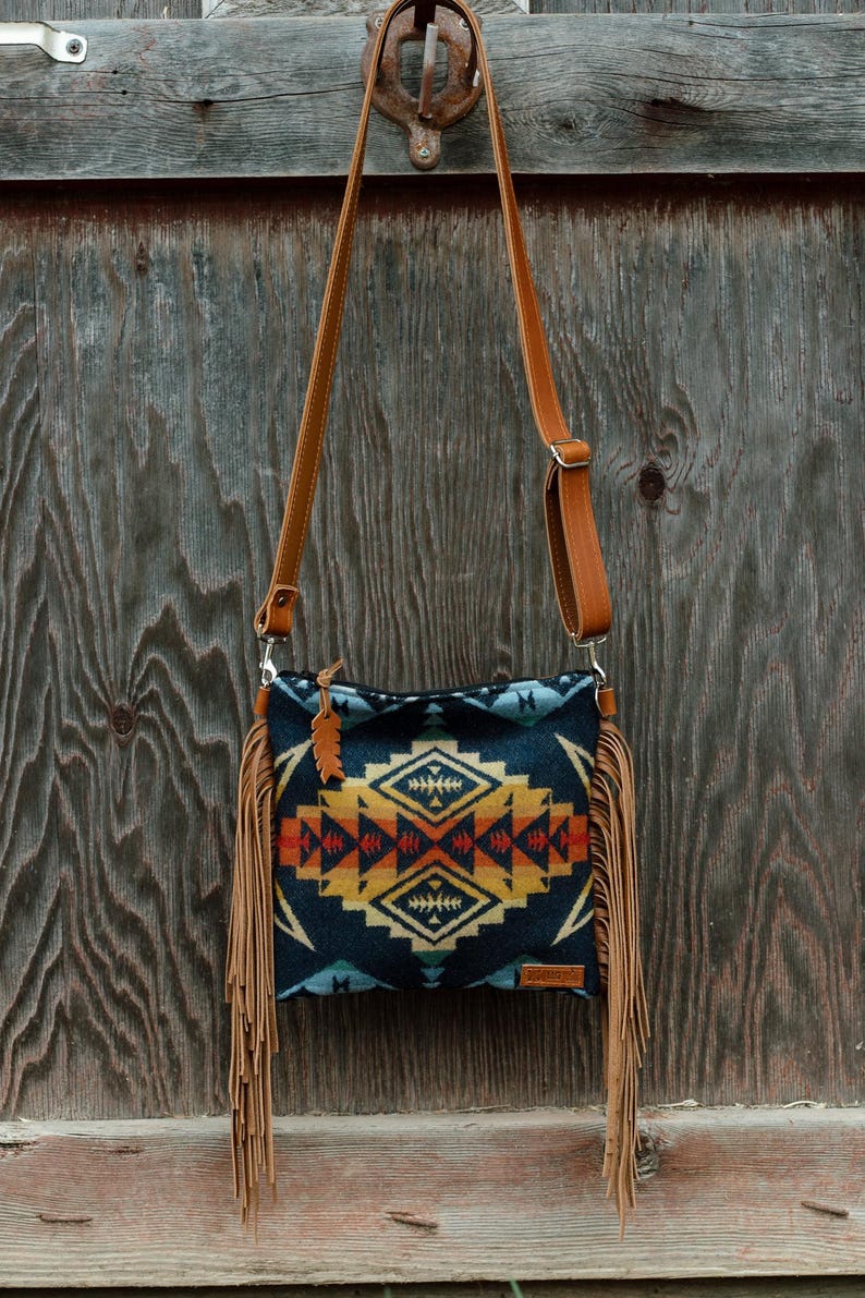 May include: A crossbody bag featuring a dark blue, orange, and yellow geometric pattern. The bag has long tan fringe on both sides, a tan adjustable shoulder strap, a zip closure with a small feather-shaped charm, and a rectangular brand tag.