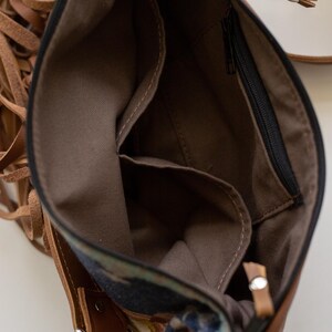 May include: A brown leather crossbody bag with long fringe accents on the side. The interior is lined with brown fabric and features an open slip pocket and a zippered pocket. The bag includes a black zipper closure and silver metal hardware.