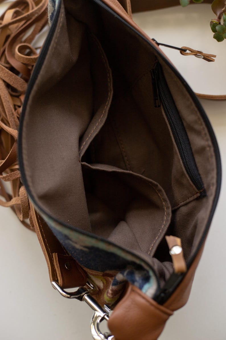 May include: A brown leather crossbody bag with long fringe accents on the side. The interior is lined with brown fabric and features an open slip pocket and a zippered pocket. The bag includes a black zipper closure and silver metal hardware.