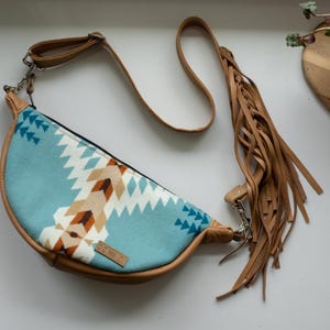 K&ouml;nnte beinhalten: Eine halbmondf&ouml;rmige Umh&auml;ngetasche mit einem hellblauen Stoffeinsatz, der ein geometrisches Muster in Wei&szlig;, Beige und Dunkelbraun aufweist. Die Tasche hat beigefarbene Kunstlederbes&auml;tze, einen verstellbaren Riemen und lange Fransen.