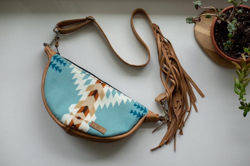 K&ouml;nnte beinhalten: Eine halbmondf&ouml;rmige Umh&auml;ngetasche mit einem hellblauen Stoffeinsatz, der ein geometrisches Muster in Wei&szlig;, Beige und Dunkelbraun aufweist. Die Tasche hat beigefarbene Kunstlederbes&auml;tze, einen verstellbaren Riemen und lange Fransen.
