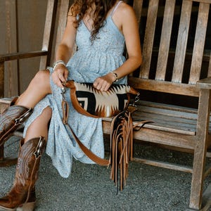 May include: A light blue floral print dress with spaghetti straps, paired with brown leather cowboy boots featuring intricate stitching and a crescent-shaped crossbody bag with a geometric pattern and long brown and black leather fringe.