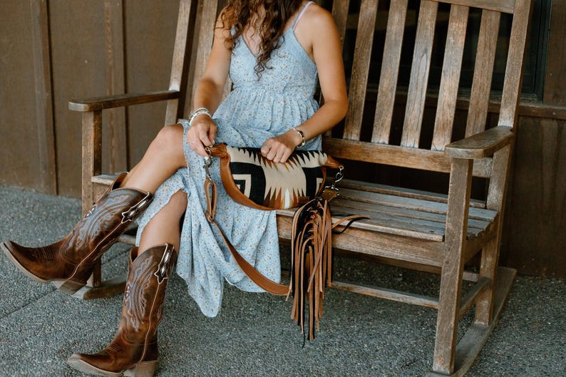 May include: A light blue floral print dress with spaghetti straps, paired with brown leather cowboy boots featuring intricate stitching and a crescent-shaped crossbody bag with a geometric pattern and long brown and black leather fringe.