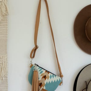 May include: A crescent-shaped crossbody bag featuring a light blue, white, and brown geometric pattern. The bag includes a long, adjustable tan strap and a large, matching tan leather-like tassel attached to a silver-toned metal clasp on the side.