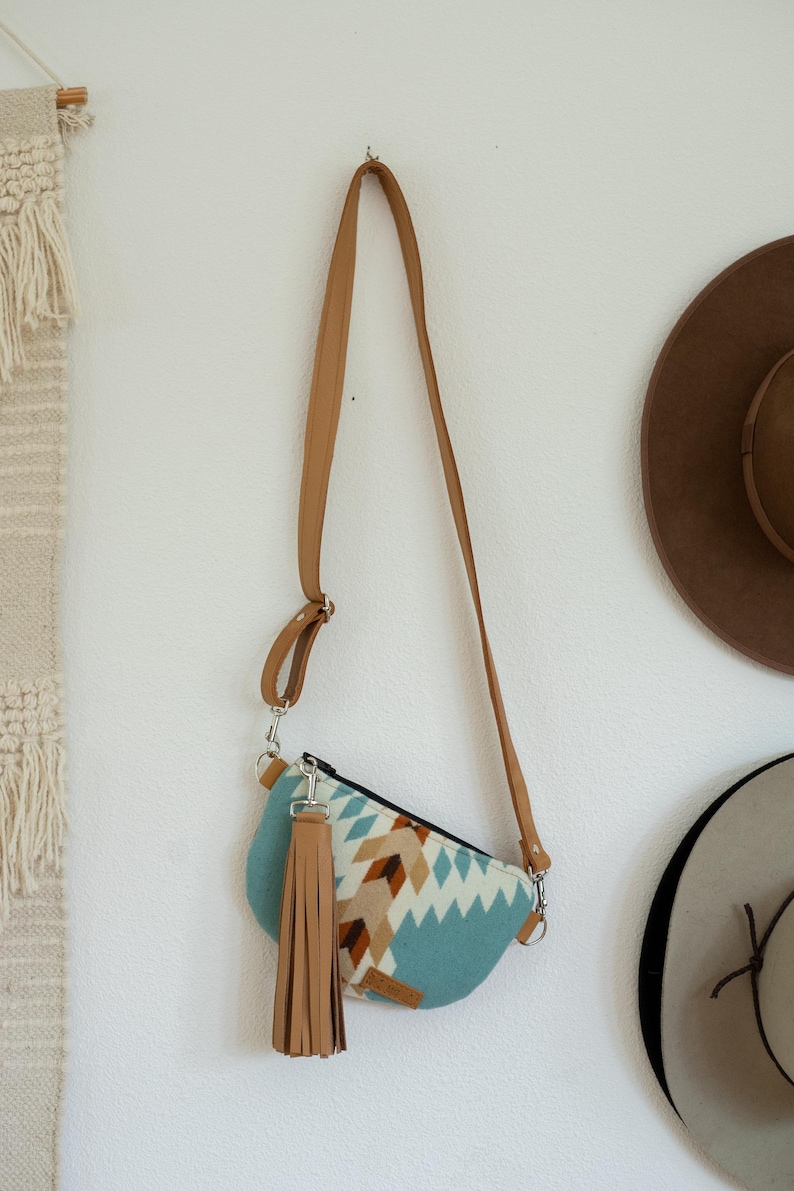 May include: A crescent-shaped crossbody bag featuring a light blue, white, and brown geometric pattern. The bag includes a long, adjustable tan strap and a large, matching tan leather-like tassel attached to a silver-toned metal clasp on the side.