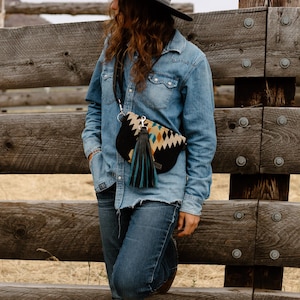 May include: A black and patterned crossbody bag with a long black strap and a teal tassel. The bag features a geometric design in tan, teal, and white. The bag is worn over a denim shirt and jeans.