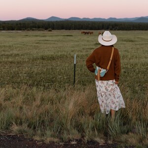 May include: A white cowboy hat, a brown long-sleeved sweater, and a white floral print midi skirt. A small light blue and white patterned crossbody bag with a tan leather strap and a long tan tassel hangs over the shoulder of the sweater.