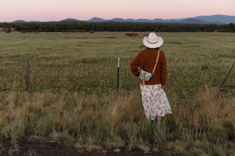 May include: A white cowboy hat, a brown long-sleeved sweater, and a white floral print midi skirt. A small light blue and white patterned crossbody bag with a tan leather strap and a long tan tassel hangs over the shoulder of the sweater.