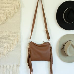 May include: A brown crossbody bag featuring long fringe detailing on both sides, a top zip closure, and an adjustable shoulder strap with metal hardware. The bag has a smooth, solid-coloured exterior and is suspended by its strap.