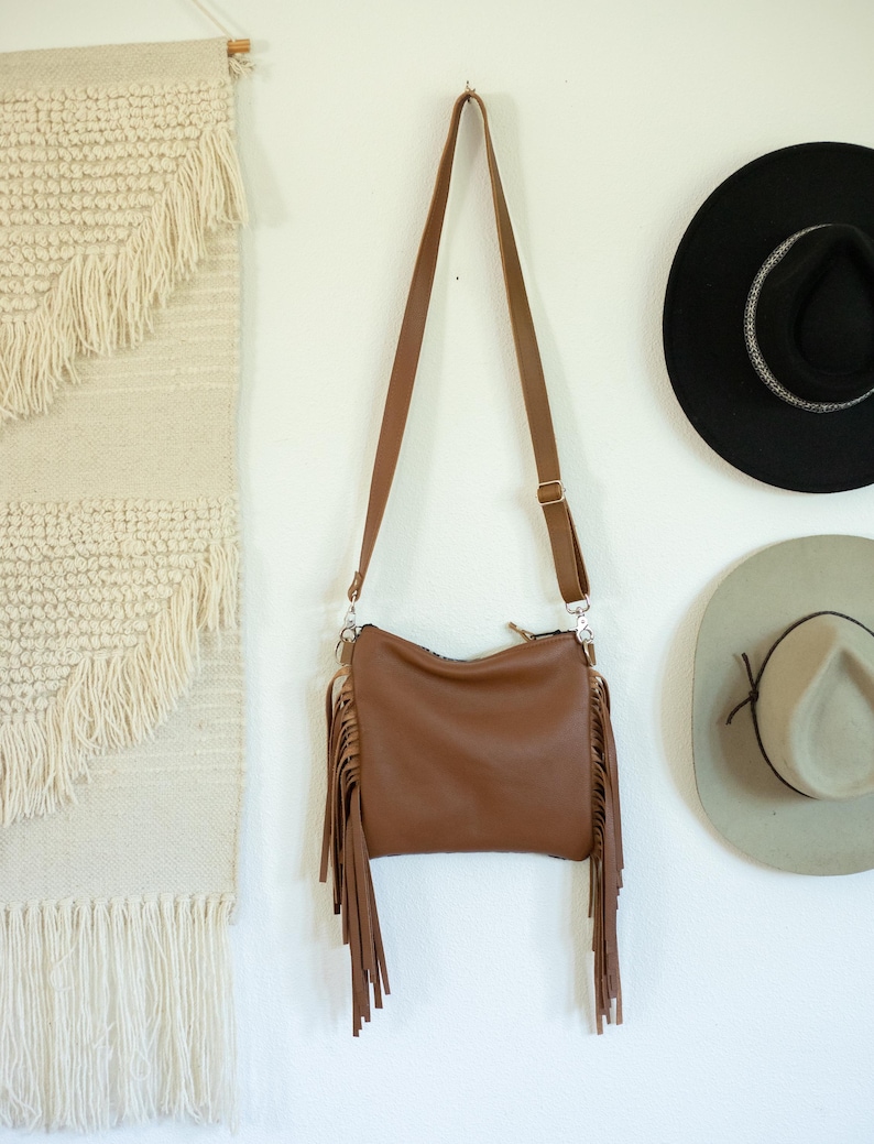 May include: A brown crossbody bag featuring long fringe detailing on both sides, a top zip closure, and an adjustable shoulder strap with metal hardware. The bag has a smooth, solid-coloured exterior and is suspended by its strap.