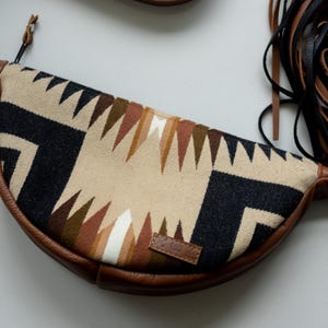 May include: A crescent-shaped crossbody bag featuring a woven fabric panel with a geometric pattern in black, cream, and shades of brown. The bag has brown leather trim, a zipper closure, and long black and brown leather fringe tassels on one side.
