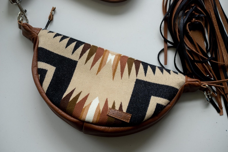 May include: A crescent-shaped crossbody bag featuring a woven fabric panel with a geometric pattern in black, cream, and shades of brown. The bag has brown leather trim, a zipper closure, and long black and brown leather fringe tassels on one side.