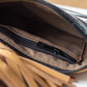 May include: An open leather pouch featuring a tan leather exterior with a long fringe detail and a black zipper closure. The interior is lined with a brown fabric and includes a smaller internal pocket with a black zipper for secure storage.