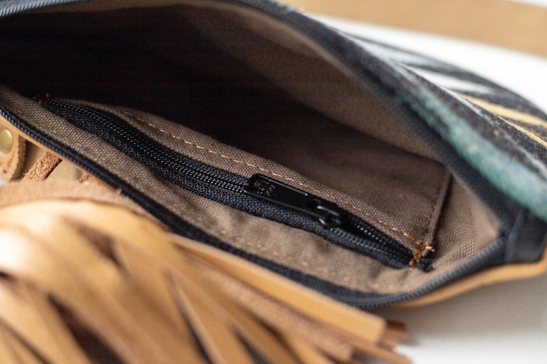 May include: An open leather pouch featuring a tan leather exterior with a long fringe detail and a black zipper closure. The interior is lined with a brown fabric and includes a smaller internal pocket with a black zipper for secure storage.