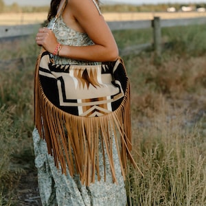 May include: A crescent-shaped shoulder bag featuring a geometric pattern in black, tan, and white. The bag is adorned with long, tan-colored fringe hanging from the bottom edge and includes a matching tan shoulder strap for carrying.