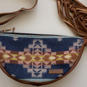 May include: A crescent-shaped fanny pack featuring a blue, brown, and cream geometric pattern on the front panel. The bag includes brown leather trim, an adjustable strap with a metal clip, and long brown leather fringe tassels on the side.