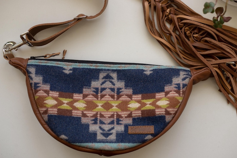 May include: A crescent-shaped fanny pack featuring a blue, brown, and cream geometric pattern on the front panel. The bag includes brown leather trim, an adjustable strap with a metal clip, and long brown leather fringe tassels on the side.