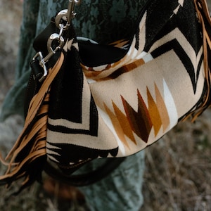May include: A patterned shoulder bag with a black, tan, and brown geometric design. The bag has a black strap and tan fringe. The bag is made of a woven material and has silver hardware.