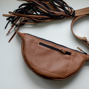 May include: A tan crescent-shaped fanny pack featuring a black zippered pocket on the front, a long adjustable strap, and a decorative fringe attachment made of alternating black and tan leather strips connected by a silver metal swivel clasp.