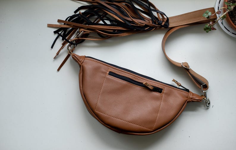 May include: A tan crescent-shaped fanny pack featuring a black zippered pocket on the front, a long adjustable strap, and a decorative fringe attachment made of alternating black and tan leather strips connected by a silver metal swivel clasp.