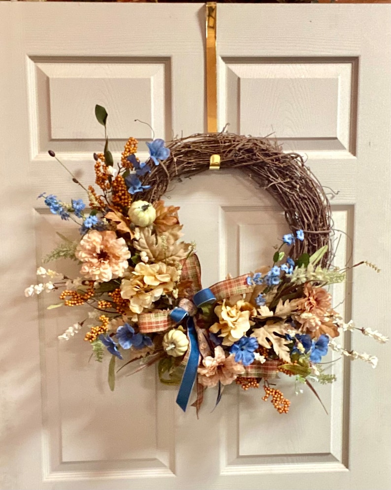 May include: A fall wreath with blue and cream colored flowers, brown vines, and a plaid ribbon. The wreath is hanging on a white door.