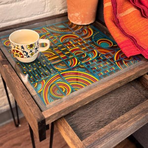May include: A wooden side table with a colorful, geometric patterned glass top and a pull-out drawer. The table has black hairpin legs and a small teacup with a floral design. A potted lavender plant and a red blanket are also visible.