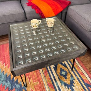 May include: Square coffee table with a dark gray wooden frame and a patterned glass top. The table has black hairpin legs and sits on a colorful rug. Two decorative mugs are on the table, and a red and orange blanket is on the sofa.