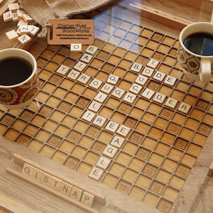 K&ouml;nnte beinhalten: Ein Holzspielbrett mit einer Glasplatte, auf dem ein fertiggespieltes Scrabble-Spiel mit den W&ouml;rtern "THROUGH", "CIDER", "TREE" und "GISTNAP" zu sehen ist. Zwei Tassen Kaffee stehen auf dem Brett, eine mit einem braunen und orangenen Muster und die andere mit einem wei&szlig;en und braunen Muster. Das Brett wurde von Charles River Woodworks hergestellt.
