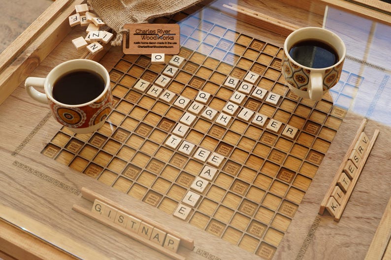 K&ouml;nnte beinhalten: Ein Holzspielbrett mit einer Glasplatte, auf dem ein fertiggespieltes Scrabble-Spiel mit den W&ouml;rtern "THROUGH", "CIDER", "TREE" und "GISTNAP" zu sehen ist. Zwei Tassen Kaffee stehen auf dem Brett, eine mit einem braunen und orangenen Muster und die andere mit einem wei&szlig;en und braunen Muster. Das Brett wurde von Charles River Woodworks hergestellt.