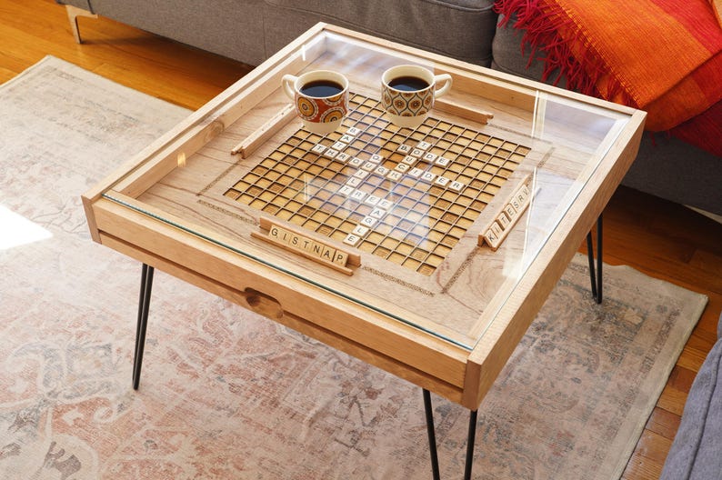 K&ouml;nnte beinhalten: Ein Holz-Couchtisch mit einer Glasplatte und einem eingebauten Scrabble-Spielbrett. Der Tisch hat schwarze Haarnadelbeine und zwei Kaffeetassen auf dem Brett.