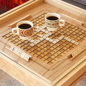 K&ouml;nnte beinhalten: Ein Holz-Couchtisch mit einer Glasplatte und einem eingebauten Scrabble-Brett. Zwei Tassen Kaffee stehen auf dem Brett, und es gibt Kacheln, die die W&ouml;rter "SEPMAIL" und "GISTNAIP" buchstabieren.